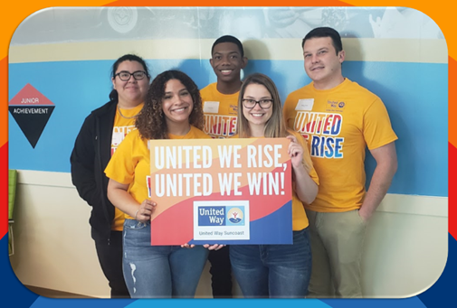 A group of five volunteers smiling and holding up a sign that says "United we rise, United we win".